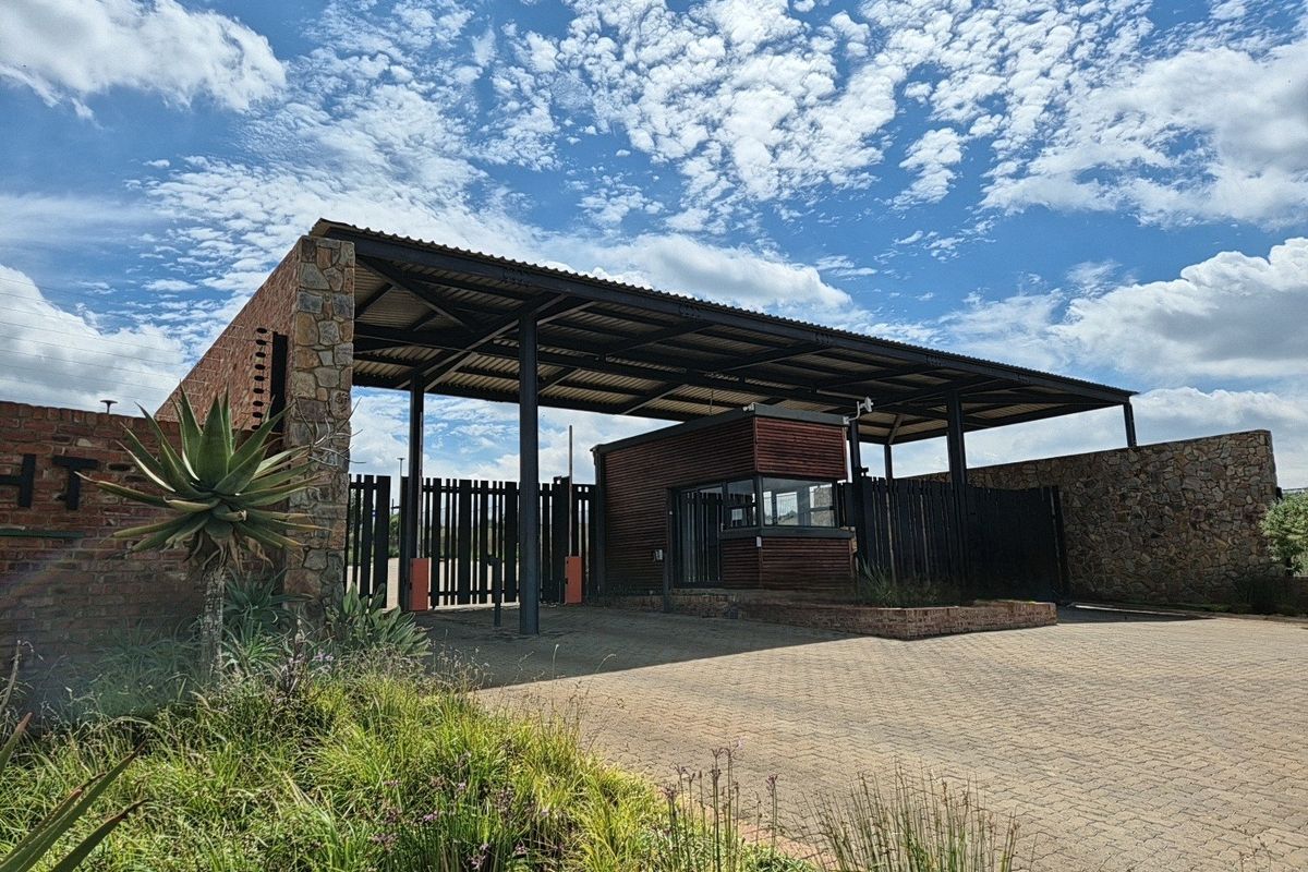 A well-maintained residential estate entrance featuring a modern security gatehouse, stone masonry walls, and automated access gates under a covered metal structure.