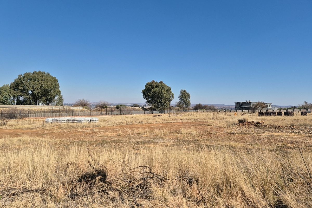 A wide-angle view of a vacant residential plot featuring dry grass, perimeter fencing, and distant landscape views.