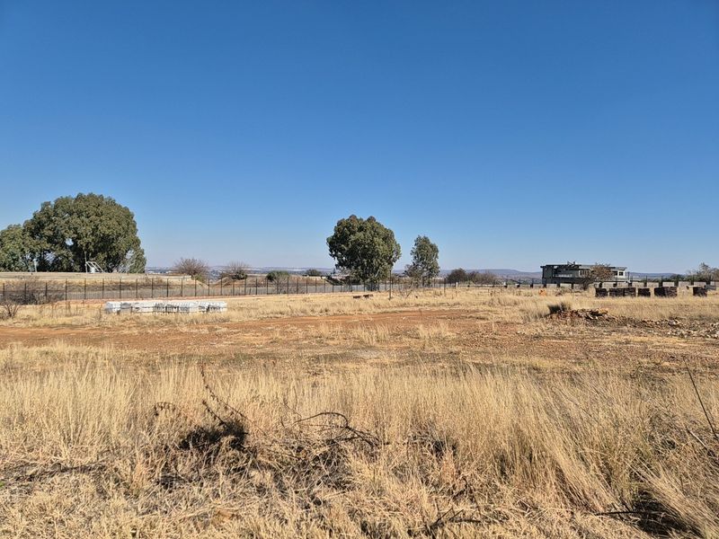 A wide-angle view of a vacant residential plot featuring dry grass, perimeter fencing, and distant landscape views.