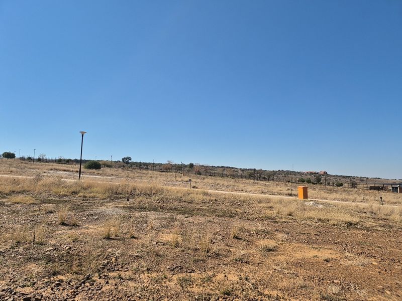A vacant residential plot featuring natural dry grass, perimeter fencing, and existing street lighting infrastructure.