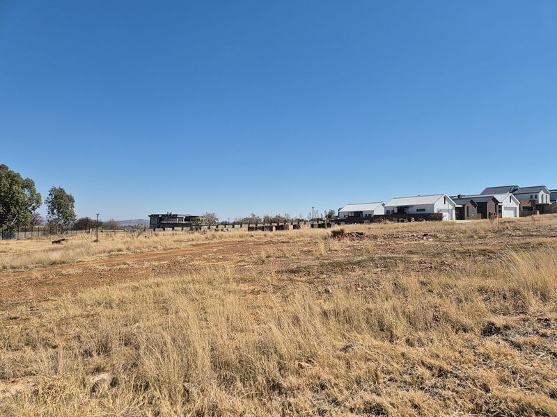 A wide-angle view of a vacant residential plot featuring dry grass and surrounding neighborhood development in the distance.