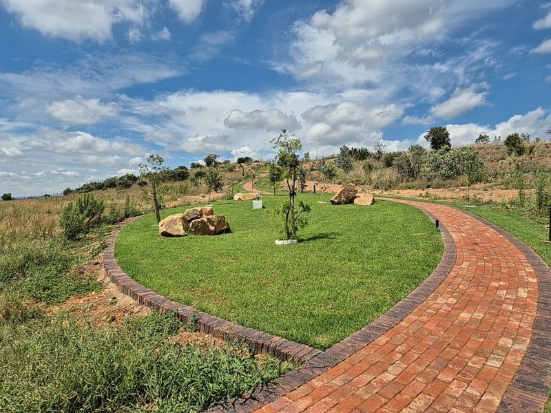 A well-maintained residential garden featuring a curved brick paved pathway, manicured lawn, and decorative landscaping elements set against a natural hillside backdrop.