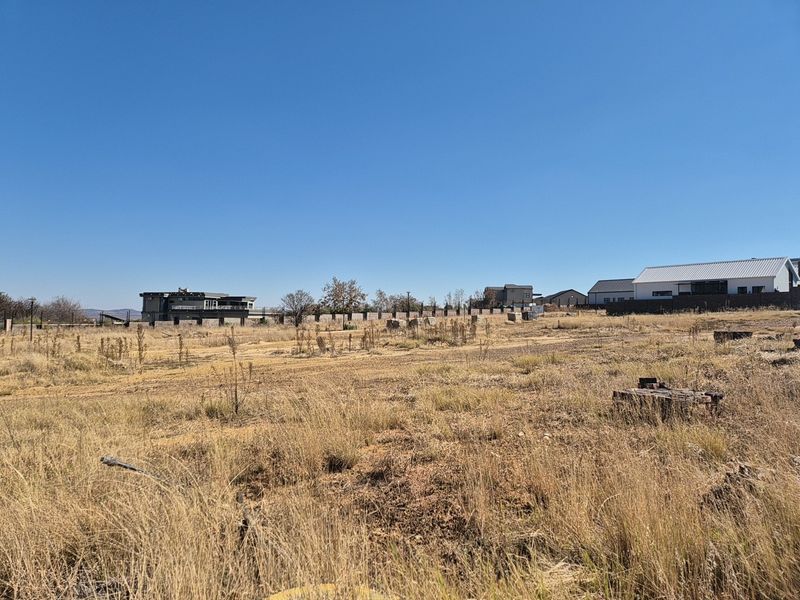 A view of a vacant residential plot featuring dry grass and perimeter fencing, situated in a developing neighborhood.