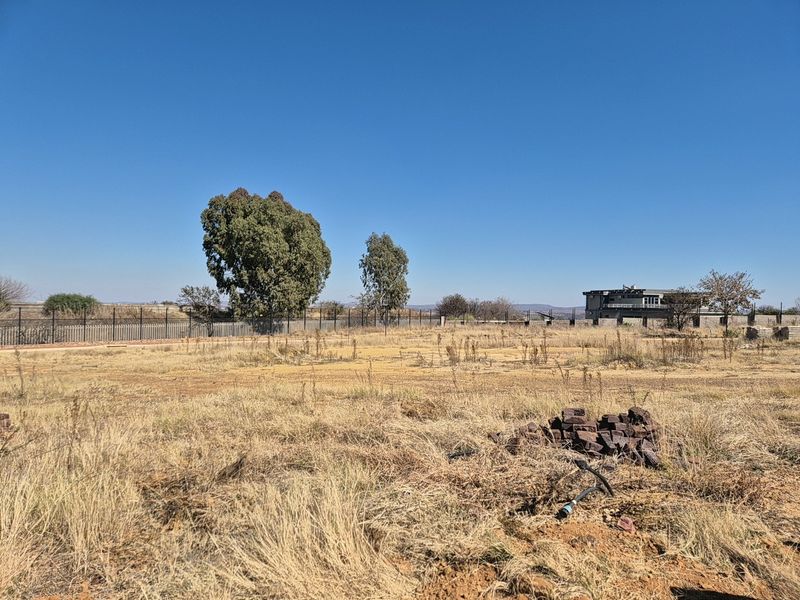 A view of a vacant residential plot featuring dry grass, perimeter fencing, and mature trees under a clear blue sky.