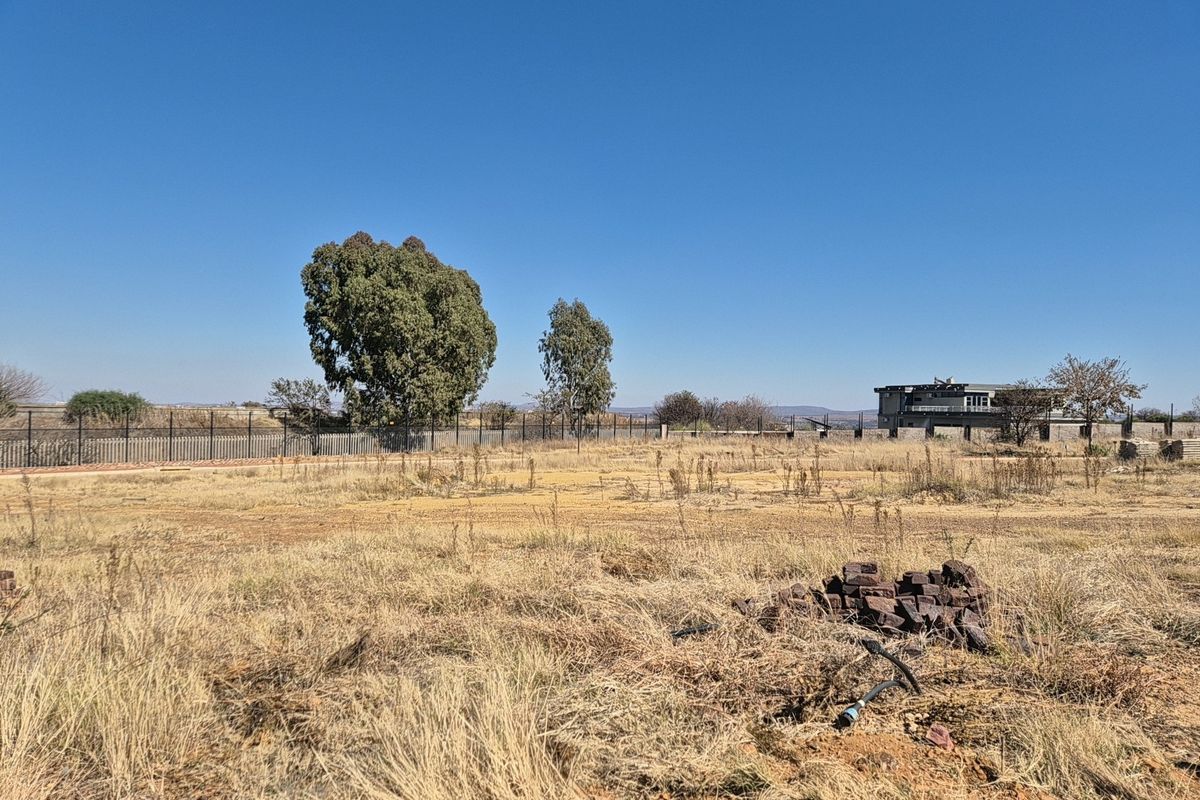 A view of a vacant residential plot featuring dry grass, perimeter fencing, and mature trees under a clear blue sky.