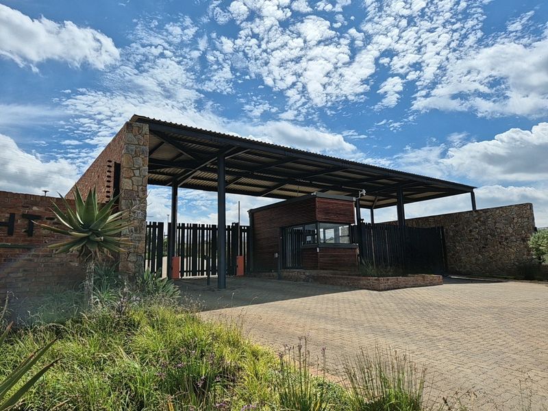 A well-maintained residential estate entrance featuring a secure guard house, stone masonry walls, paved driveway, and robust steel gate infrastructure.