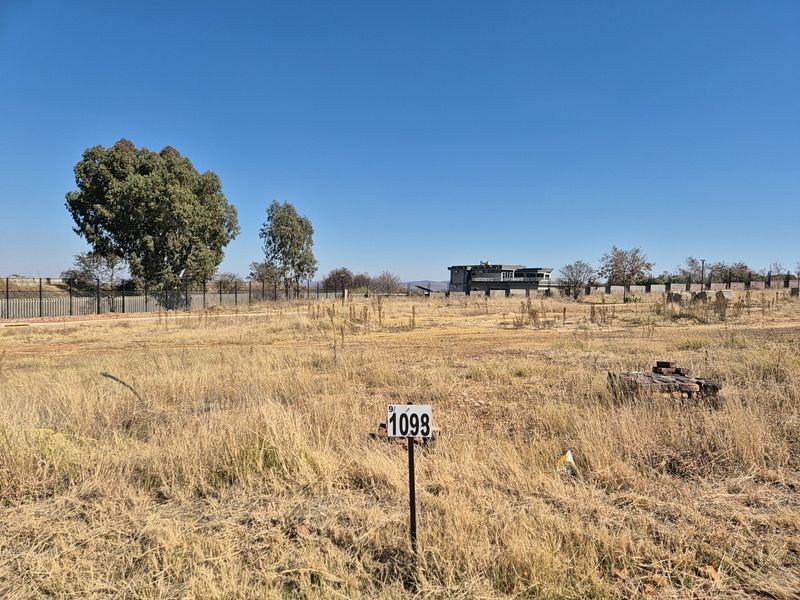 A vacant residential plot featuring dry grass, perimeter fencing, and a clear lot marker, ready for development.