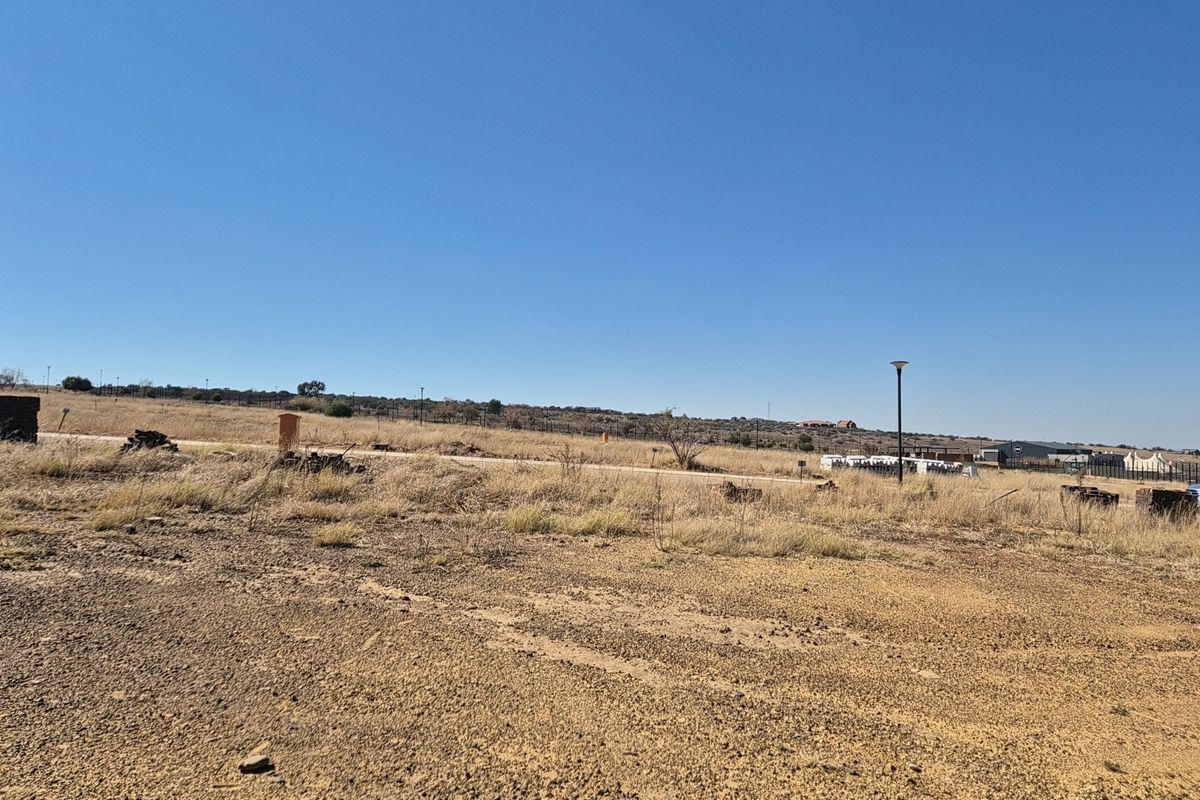 A vacant residential plot featuring dry natural vegetation, street lighting, and perimeter fencing, offering a clear view of the surrounding landscape.