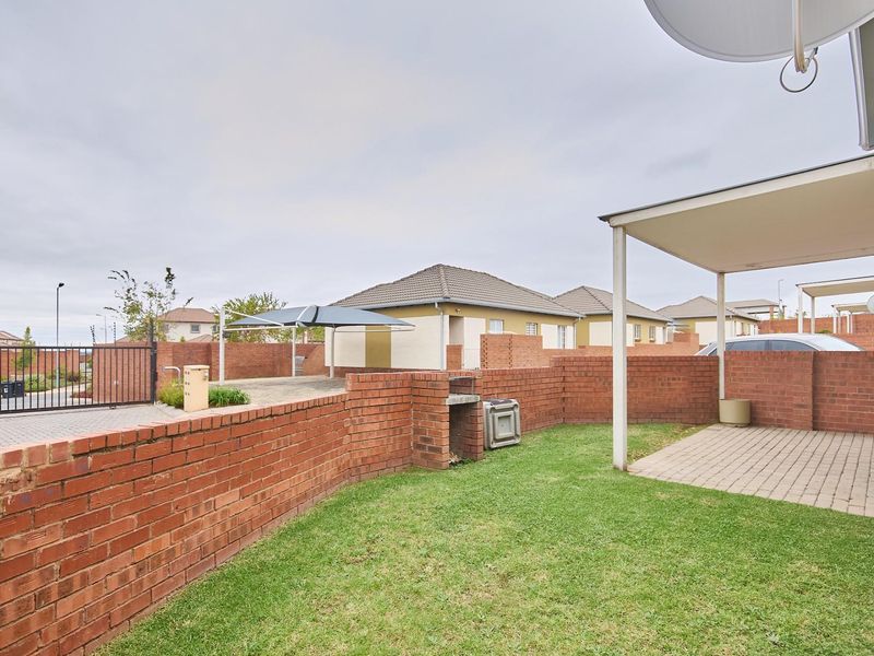 A private rear garden area featuring a brick boundary wall, a built-in braai, a paved patio section, and a covered carport structure.