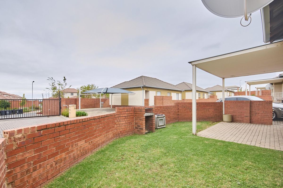 A private rear garden area featuring a brick boundary wall, a built-in braai, a paved patio section, and a covered carport structure.