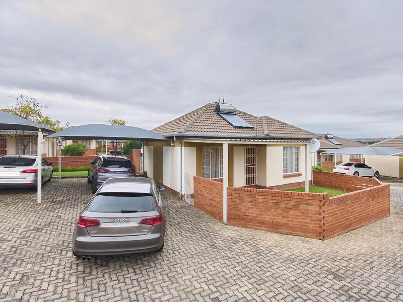 Front view of a single-story residential house featuring a paved driveway, covered carport, brick boundary wall, and a solar water heating system.