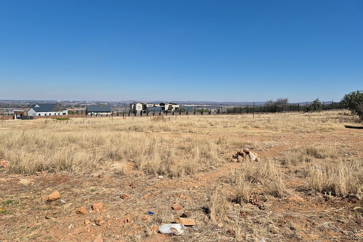 A vacant residential plot featuring dry grass and open land with distant views of neighboring estate properties.