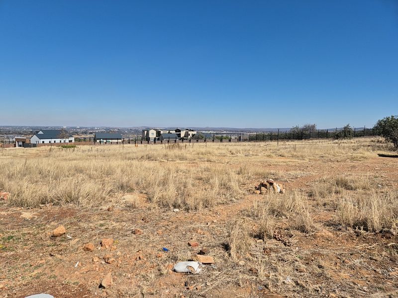 A vacant residential plot featuring dry grass and open land with distant views of neighboring estate properties.