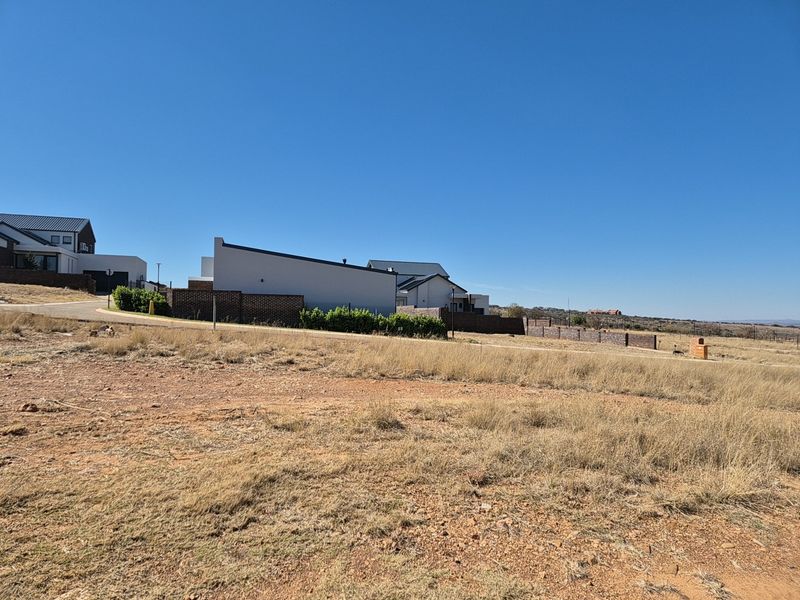 A view of a vacant residential plot showing the surrounding neighborhood, paved road access, and boundary fencing in a clear, sunny setting.