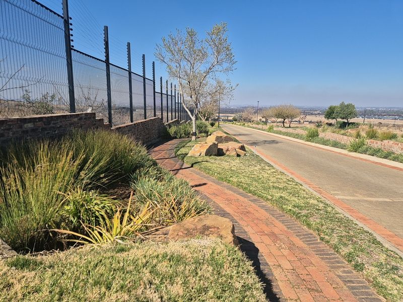 A well-maintained residential street view featuring a paved walkway, brick retaining wall, electric fencing, and landscaped garden areas.