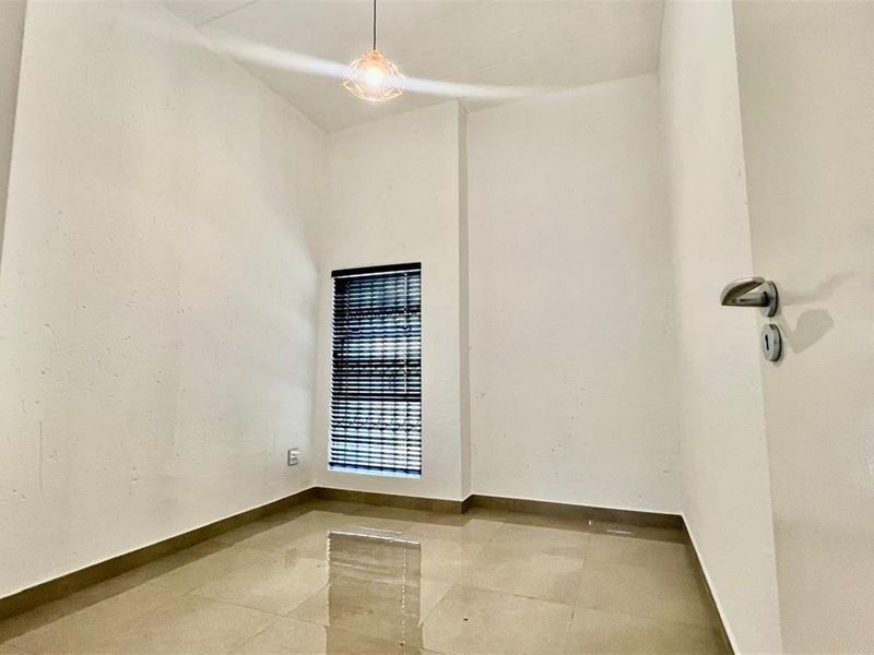 A bright, empty bedroom featuring neutral tiled flooring, clean white walls, and a window fitted with venetian blinds.