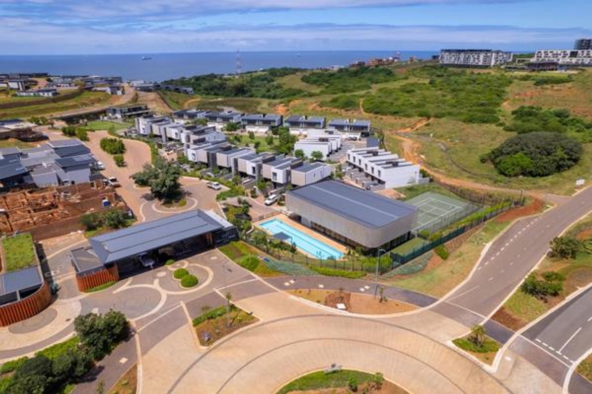 Aerial view of a modern residential estate featuring a clubhouse, swimming pool, tennis court, and surrounding landscaped grounds with coastal views.