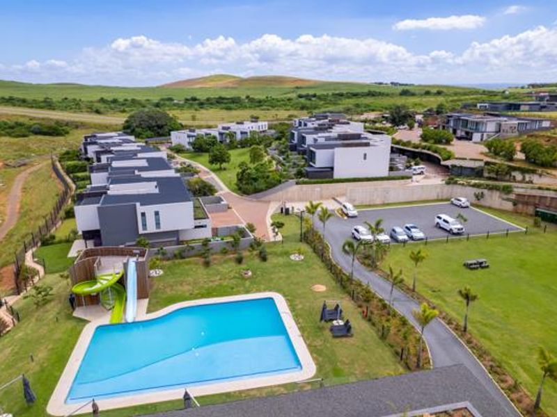 Aerial view of a residential estate featuring modern townhouses, a communal swimming pool with a slide, landscaped gardens, and paved parking areas.