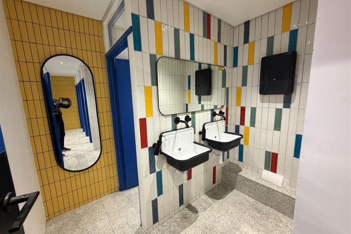Modern office restroom featuring dual wall-mounted industrial-style sinks, vibrant multi-colored tiled walls, and terrazzo-style flooring.