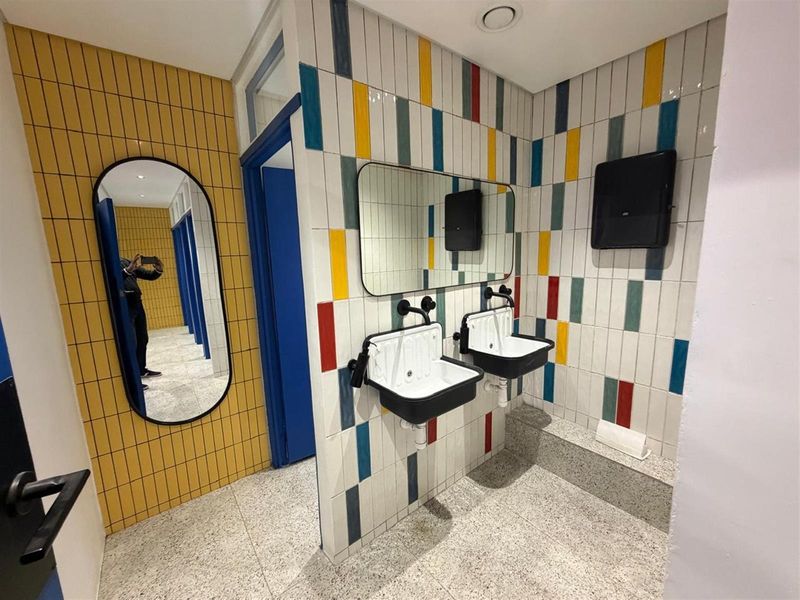 Modern office restroom featuring dual wall-mounted industrial-style sinks, vibrant multi-colored tiled walls, and terrazzo-style flooring.