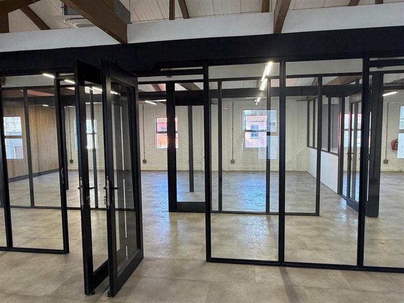 Modern open-plan office space featuring industrial-style black metal and glass partitions, exposed timber ceiling beams, and polished concrete flooring.