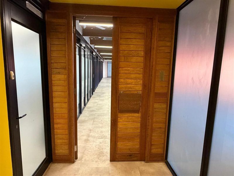 A view through a wooden door into a long office corridor featuring tiled flooring and glass-partitioned office spaces.