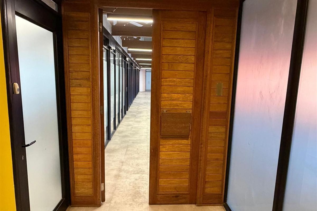 A view through a wooden door into a long office corridor featuring tiled flooring and glass-partitioned office spaces.