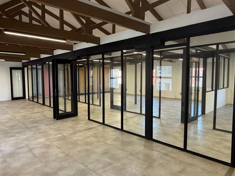 Modern open-plan office space featuring exposed timber roof trusses, polished tile flooring, and sleek black-framed glass partitioning for private meeting rooms.