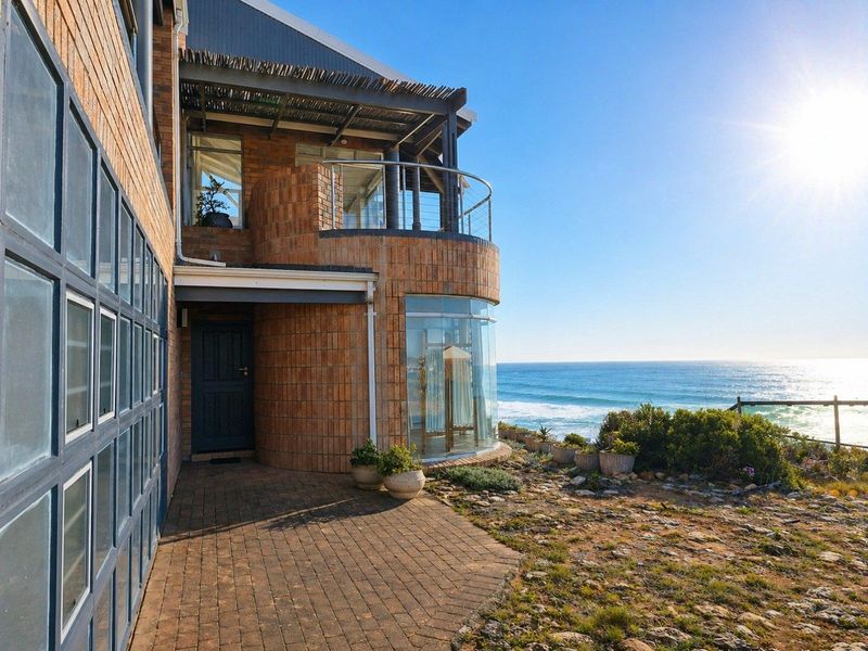 A coastal home featuring a brick facade, curved glass windows, a covered balcony with a thatched roof, and direct ocean views.