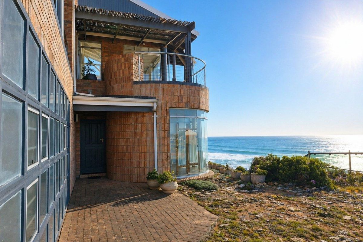 A coastal home featuring a brick facade, curved glass windows, a covered balcony with a thatched roof, and direct ocean views.