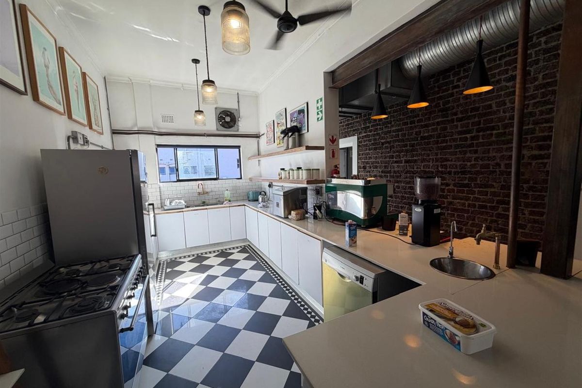 A well-lit office kitchen featuring a checkerboard floor, exposed brick wall, industrial-style pendant lighting, and white cabinetry with stone countertops.