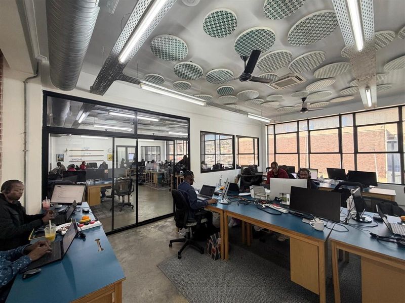 Modern open-plan office space featuring industrial-style exposed ducting, acoustic ceiling panels, large windows, and glass-partitioned meeting areas.