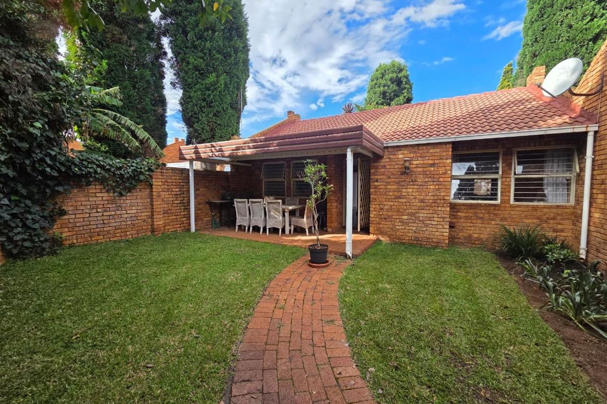 A private rear garden featuring a brick-paved pathway leading to a covered patio area, surrounded by established greenery and brick boundary walls.