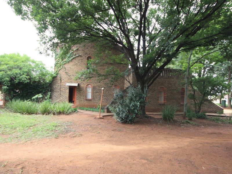 A unique face brick house featuring arched windows and security bars, set within a large, mature garden with an unpaved driveway.