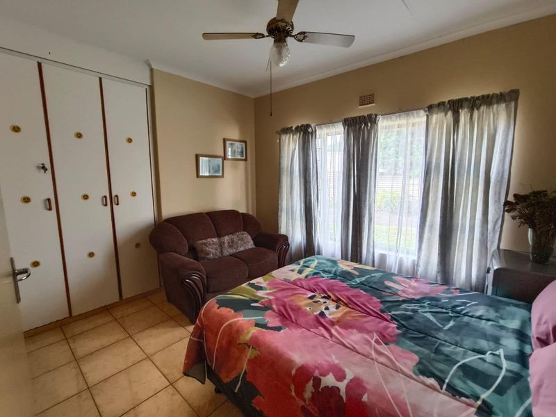 A bedroom featuring tiled flooring, built-in cupboards, and a ceiling fan, offering a functional space with natural light through large windows.