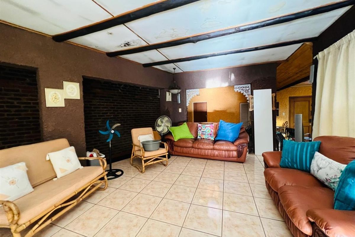 A living room featuring tiled flooring, exposed ceiling beams, and a textured brick accent wall, showing signs of wear and ceiling water damage.