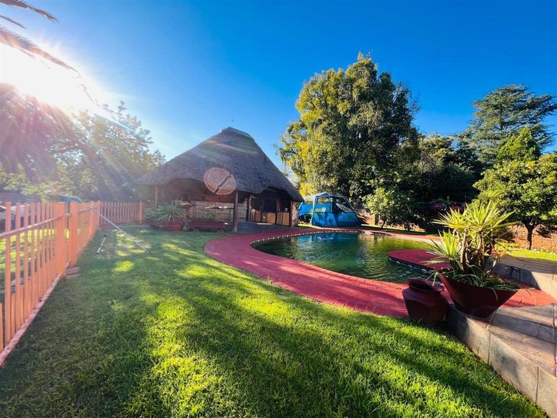 Outdoor pool area featuring a swimming pool with a red paved surround and a thatched lapa structure in a landscaped garden setting.