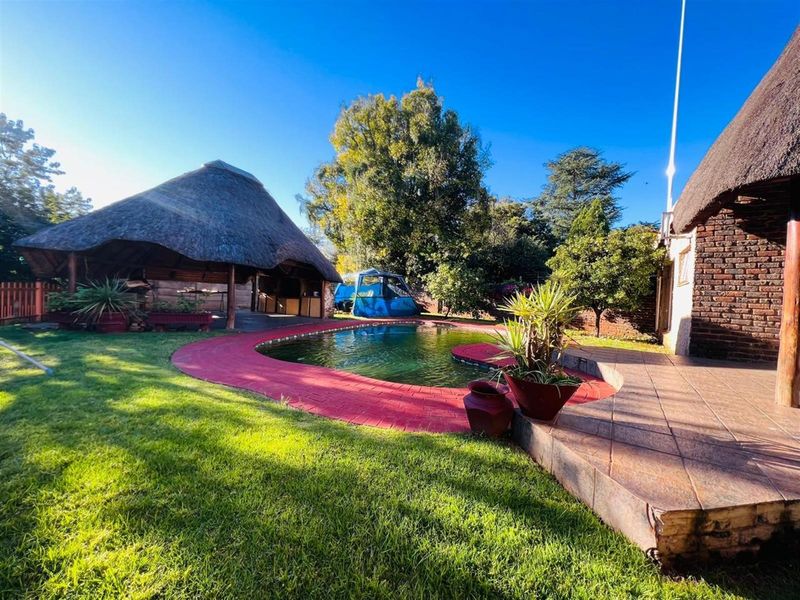 Outdoor pool area featuring a swimming pool with red brick coping, a thatched lapa entertainment area, and surrounding garden space.