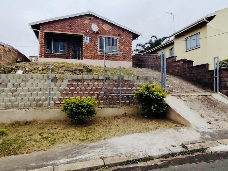 Front view of a single-story brick house featuring a paved driveway, retaining wall, and security fencing.