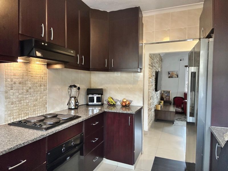 A functional kitchen featuring dark wood cabinetry, granite countertops, a built-in electric hob, extractor fan, and tiled backsplash, leading into an adjacent living area.