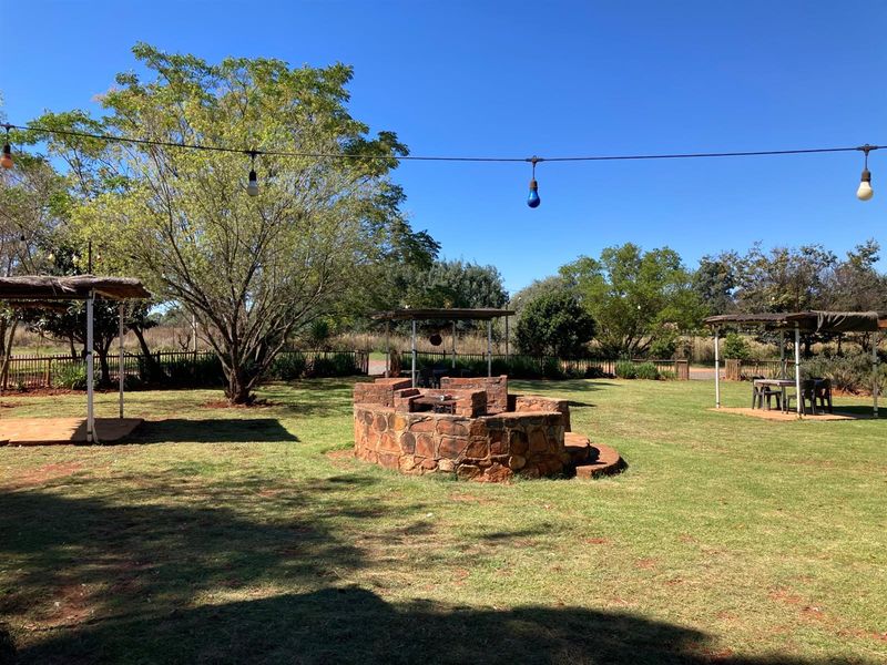 Spacious outdoor garden area featuring a central stone-built braai facility, multiple shaded seating structures, and overhead string lighting.