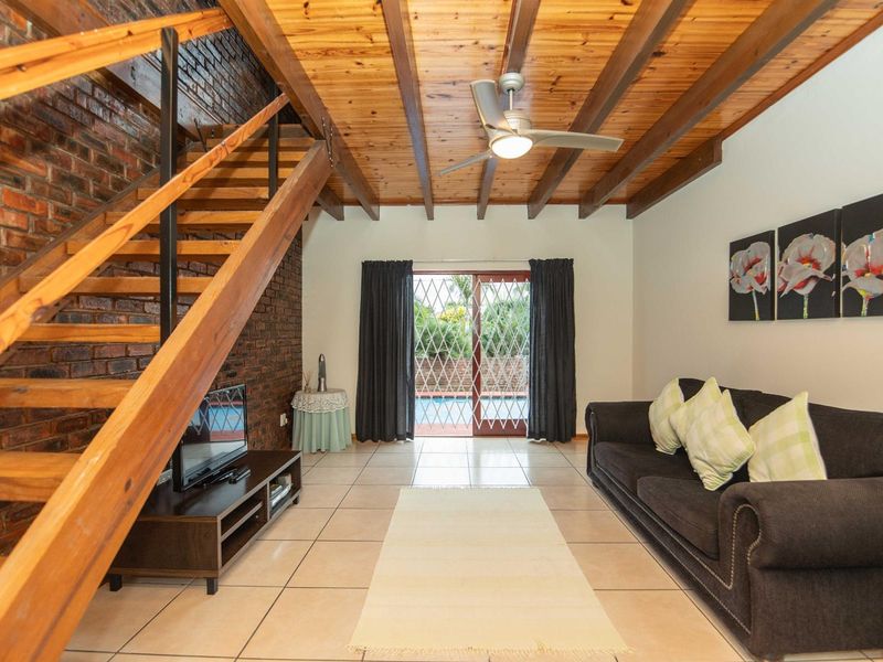 Spacious living room featuring a timber-beamed ceiling, exposed brick feature wall, staircase, and sliding glass doors leading to the outdoor pool area.