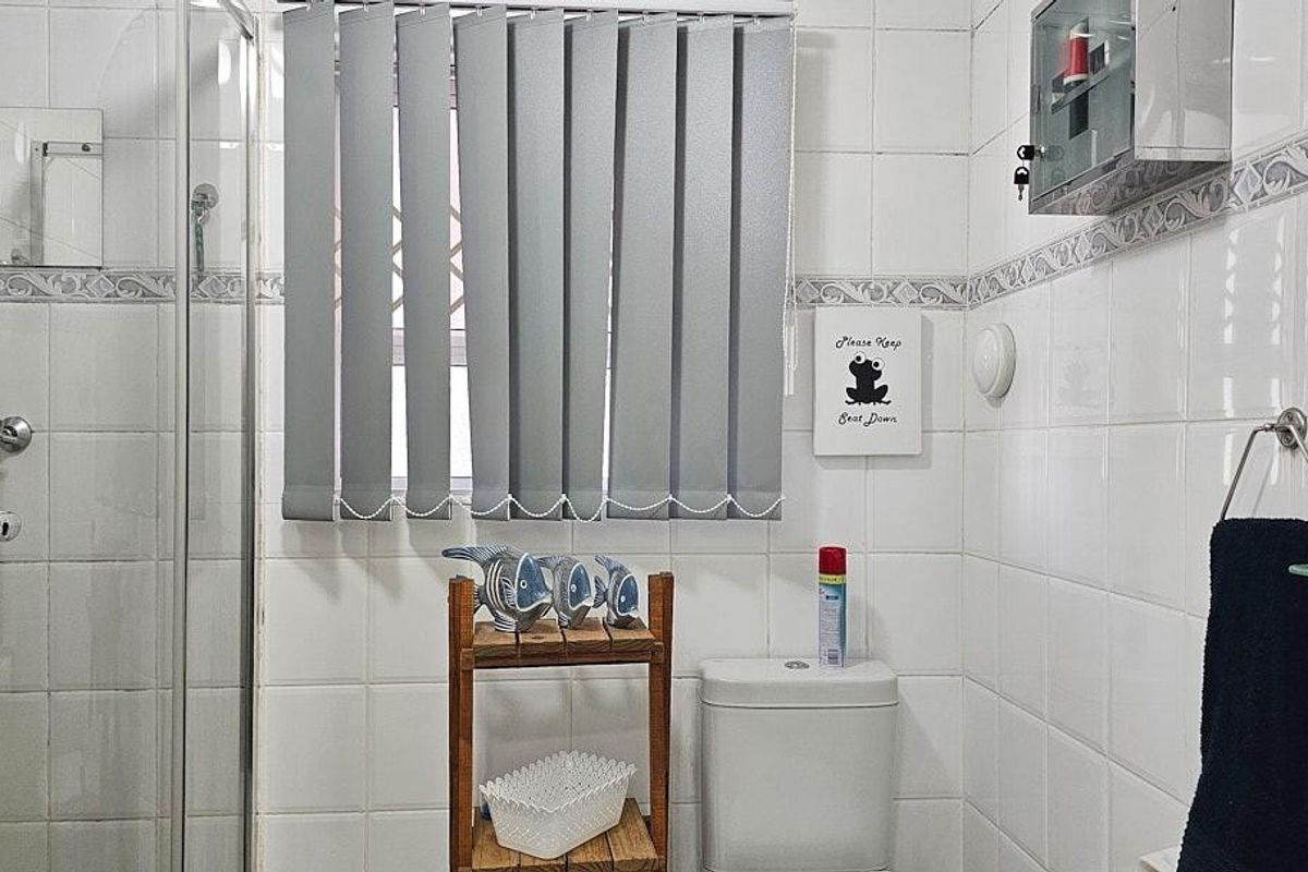 A functional bathroom featuring a glass shower enclosure, white wall tiling, and a standard toilet with vertical window blinds.