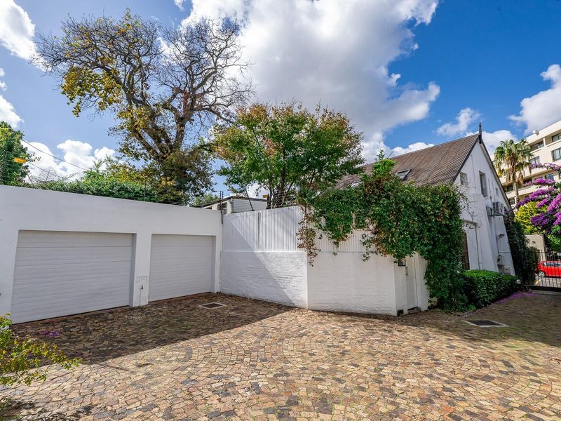 A view of the property's exterior front, featuring a double garage, paved driveway, and white perimeter walls with established greenery.