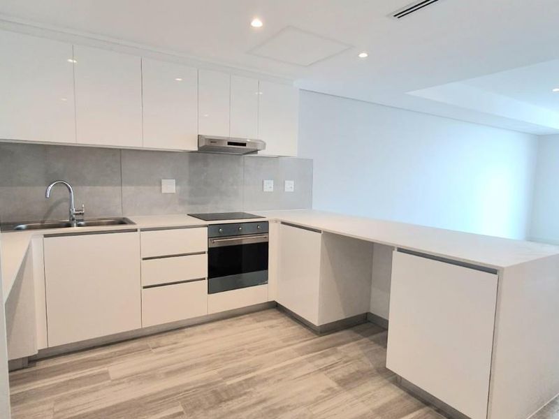 Modern kitchen featuring sleek white cabinetry, stone countertops, integrated oven, hob, extractor fan, and durable wood-look flooring in an open-plan layout.