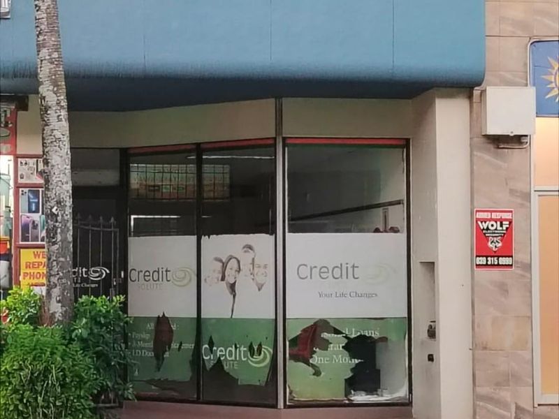 Commercial storefront entrance featuring large glass display windows with damaged signage and a blue awning, located on a paved street frontage.