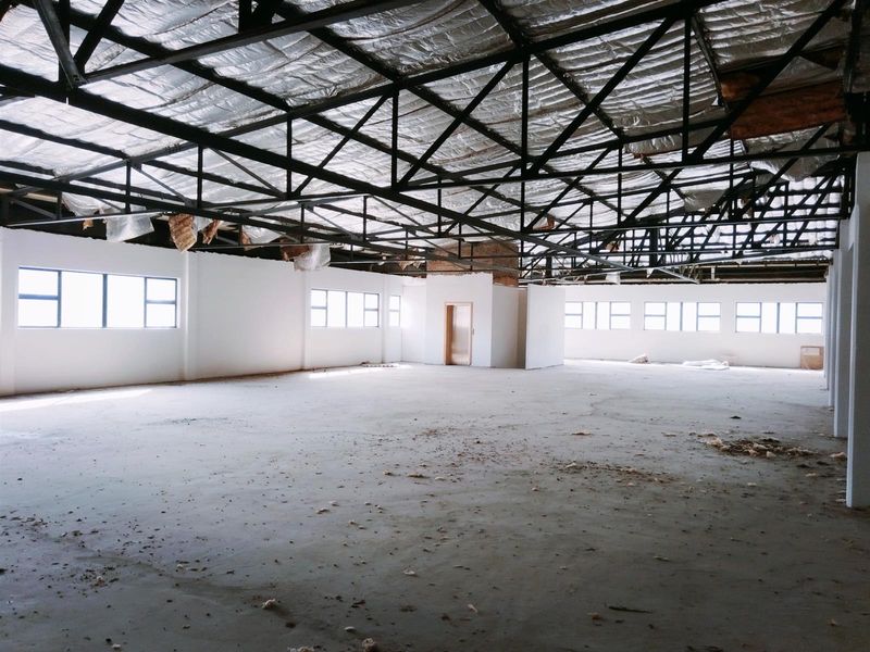A large, open-plan commercial space featuring an exposed steel truss ceiling, concrete flooring, and multiple windows providing natural light.
