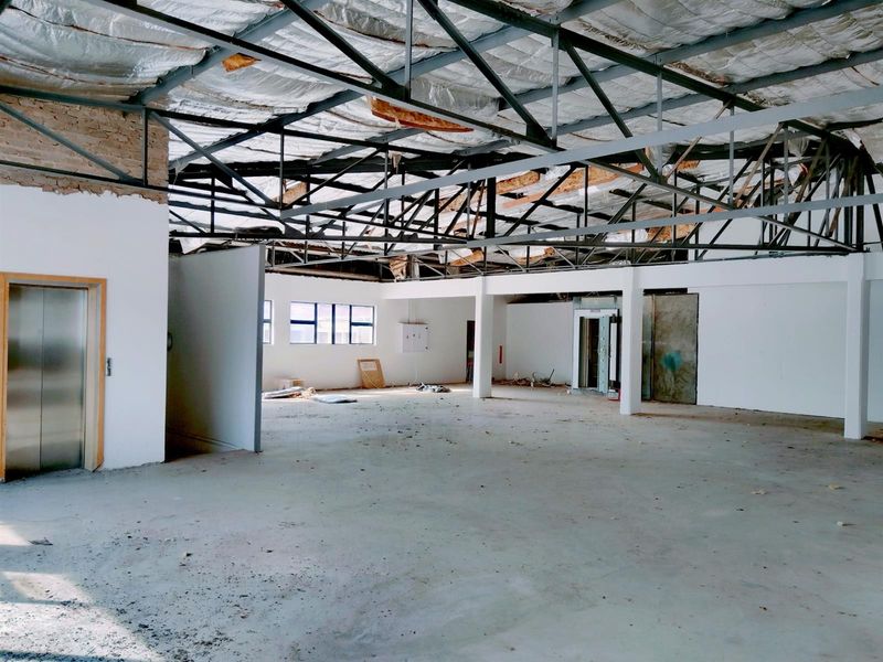 A large, open-plan commercial space currently undergoing renovation, featuring exposed steel roof trusses, concrete flooring, and an integrated elevator access point.