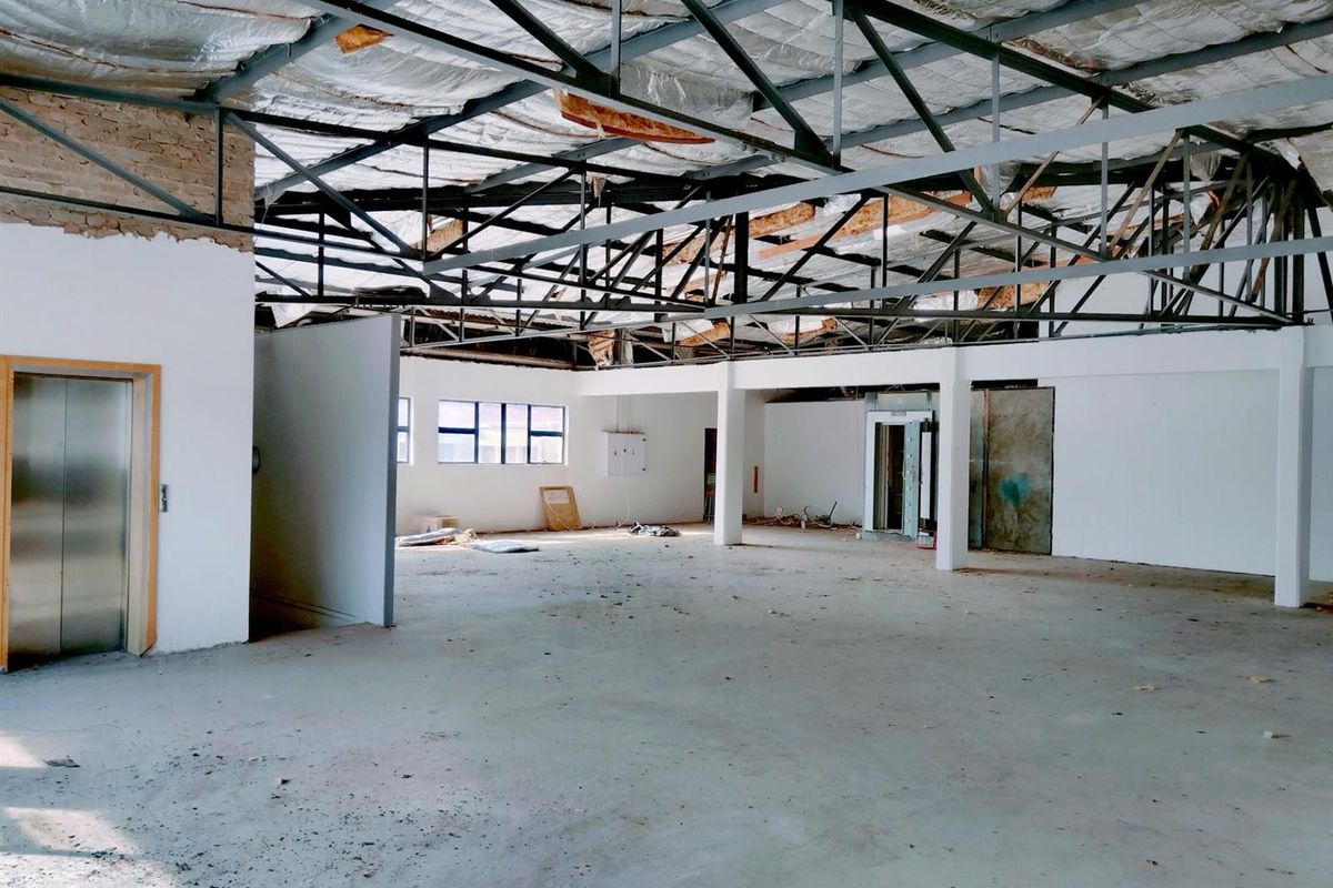 A large, open-plan commercial space currently undergoing renovation, featuring exposed steel roof trusses, concrete flooring, and an integrated elevator access point.