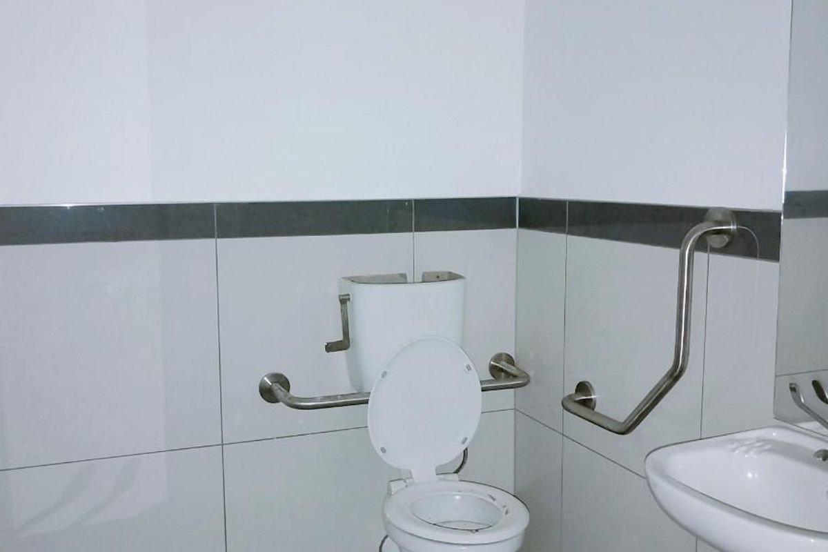 Accessible commercial restroom featuring white wall tiles with a dark accent border, stainless steel grab rails, a wall-mounted basin, and a standard toilet.